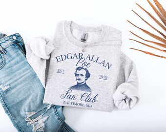 May include: A light gray sweatshirt with the text "Edgar Allan Poe Fan Club" in navy blue. The sweatshirt features a portrait of Edgar Allan Poe and the year 1809. Light blue denim jeans are visible on the left side of the image.