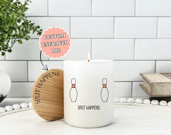 May include: A white candle with a lit wick, featuring two bowling pin illustrations and the text "SPLIT HAPPENS." The candle has a wooden lid with the same text. A pink sticker reads "CUSTOM ENGRAVED LID!"