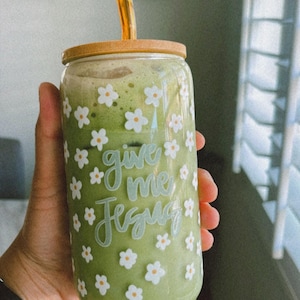 May include: A green smoothie in a glass jar with a cork lid and a gold straw. The jar has a white floral pattern and the text "give me Jesus" written on it.