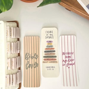 May include: Three decorative bookmarks with inspirational quotes. One bookmark has vertical peach and gray stripes and the words "Bible Cover". Another bookmark has a list of virtues and the text "Fruit of the Spirit". The third bookmark has pink and white stripes and the text "In Christ Alone my hope is found".