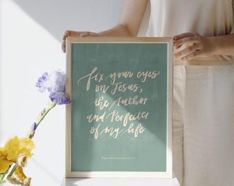 May include: A framed print with the text "fix your eyes on Jesus, the Author and Perfecter of my life" in elegant calligraphy on a teal background. The frame is light wood. A bouquet of yellow and purple flowers is in the foreground.