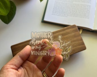 May include: A clear sticker with the text "Motherhood is my Ministry" in white script, adorned with floral illustrations in pink, orange, and green. The sticker is held in a hand, with a wooden tray and open book in the background.