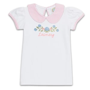 Puede incluir: Camiseta blanca de manga corta con cuello y ribete de cuadros rosas. La camiseta presenta un diseño floral bordado con el nombre "Lainey" en escritura rosa. Las mangas son abullonadas.