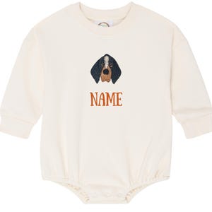 May include: A cream-coloured baby onesie with long sleeves. It features an embroidered dog face with black, brown, and white details. The word "NAME" is printed in orange below the dog's face.