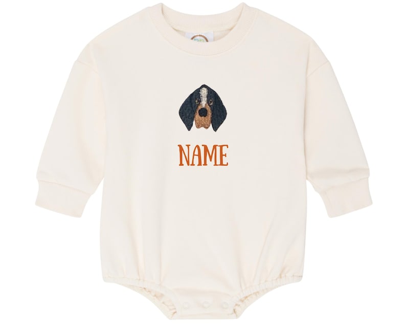May include: A cream-coloured baby onesie with long sleeves. It features an embroidered dog face with black, brown, and white details. The word "NAME" is printed in orange below the dog's face.