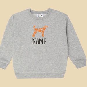 May include: A light grey sweatshirt with an orange plaid dog design and the word "NAME" in black. The dog graphic is a silhouette with an orange and white plaid pattern. The sweatshirt has a crew neck and long sleeves.