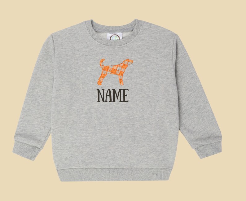 May include: A light grey sweatshirt with an orange plaid dog design and the word "NAME" in black. The dog graphic is a silhouette with an orange and white plaid pattern. The sweatshirt has a crew neck and long sleeves.