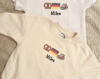 Puede incluir: Dos conjuntos para bebés, una camiseta blanca y un body color crema. Ambos presentan un diseño bordado de pretzel, bandera alemana y sombrero, con el nombre "Miles" debajo. Los conjuntos son para bebés.