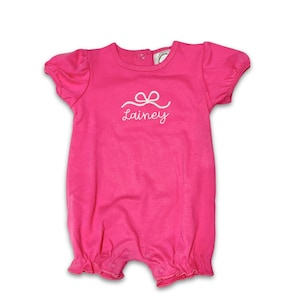 May include: A bright pink baby romper with short sleeves and ruffled leg openings. The romper features a white embroidered bow and the name "Lainey" in script. The garment has a round neckline and snap closures.