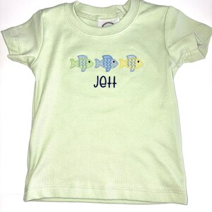 May include: A light green t-shirt with three embroidered fish in blue and yellow. The name "Jett" is embroidered in navy blue below the fish. The shirt has short sleeves and a crew neck.