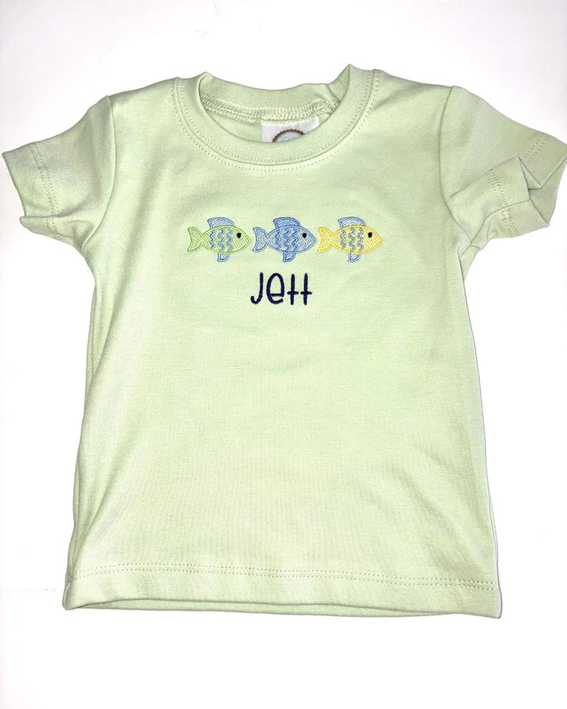 May include: A light green t-shirt with three embroidered fish in blue and yellow. The name "Jett" is embroidered in navy blue below the fish. The shirt has short sleeves and a crew neck.