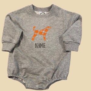 May include: A grey baby romper with long sleeves and a round neck. An orange plaid dog graphic is embroidered on the front, with the word "NAME" below it in black letters. The romper has snap closures at the bottom.