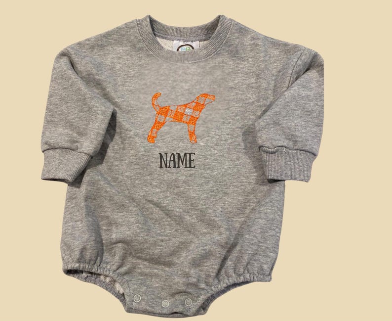 May include: A grey baby romper with long sleeves and a round neck. An orange plaid dog graphic is embroidered on the front, with the word "NAME" below it in black letters. The romper has snap closures at the bottom.
