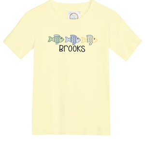 May include: A light yellow t-shirt featuring three embroidered fish in shades of green, blue, and yellow. The name "BROOKS" is embroidered below the fish. The shirt has short sleeves and a crew neck.