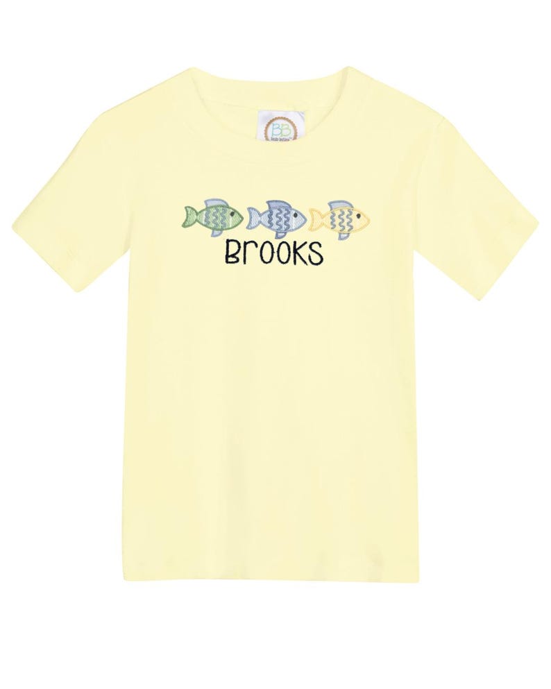 May include: A light yellow t-shirt featuring three embroidered fish in shades of green, blue, and yellow. The name "BROOKS" is embroidered below the fish. The shirt has short sleeves and a crew neck.
