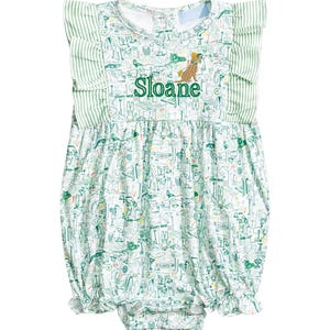 May include: White baby romper with a green golf-themed pattern and green striped ruffle sleeves. The name "Sloane" is embroidered in green, along with a cartoon animal wearing a golf visor. The romper has a bubble silhouette.