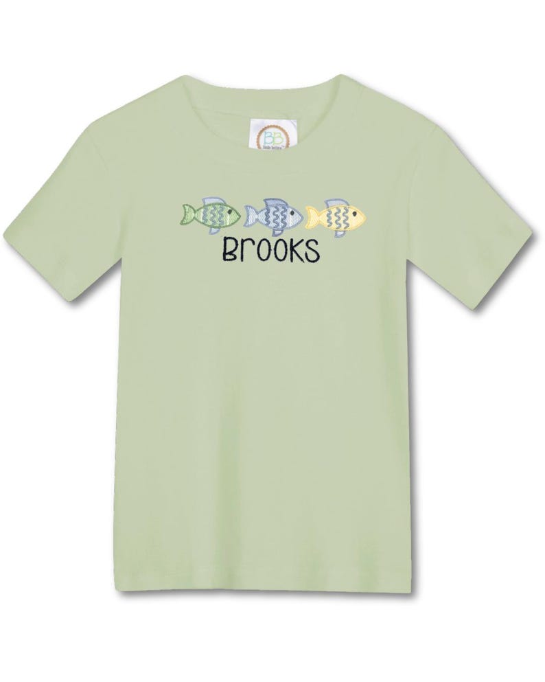 May include: A light green short-sleeved t-shirt featuring three embroidered fish in shades of green, blue, and yellow. The name "BROOKS" is embroidered below the fish. The shirt is made of a soft material.