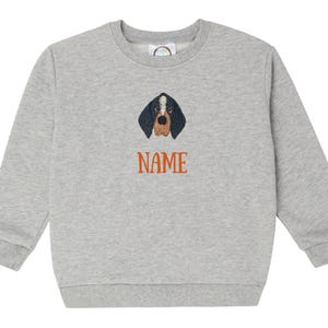 May include: A grey sweatshirt with a crew neck. It features an embroidered dog face with black and brown details. The word "NAME" is embroidered in orange below the dog's face. The sweatshirt is made of a soft material, suitable for casual wear.