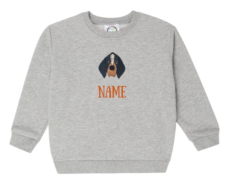 May include: A grey sweatshirt with a crew neck. It features an embroidered dog face with black and brown details. The word "NAME" is embroidered in orange below the dog's face. The sweatshirt is made of a soft material, suitable for casual wear.