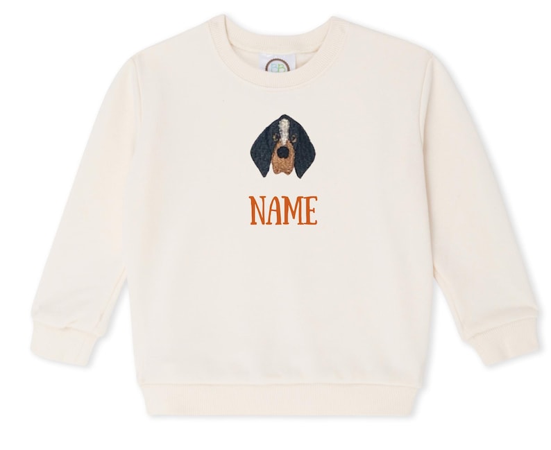 May include: Cream-coloured sweatshirt with an embroidered dog face in black and brown. The word "NAME" is embroidered in orange beneath the dog's head. The sweatshirt has a crew neck and long sleeves.