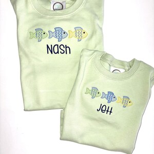 May include: Two light green shirts with embroidered fish designs. One shirt says "Nash" in navy blue, and the other says "Jett". Each shirt has three fish in green, blue, and yellow.