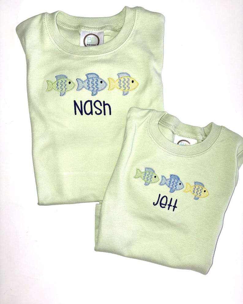 May include: Two light green shirts with embroidered fish designs. One shirt says "Nash" in navy blue, and the other says "Jett". Each shirt has three fish in green, blue, and yellow.