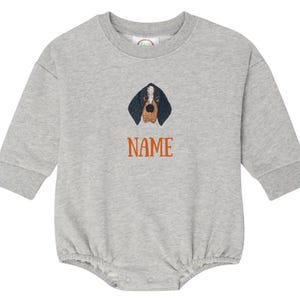 May include: A pale grey baby romper with long sleeves and a round neckline. The romper has an embroidered dog face with black, brown, and white details. The word "NAME" is embroidered in orange below the dog's face.
