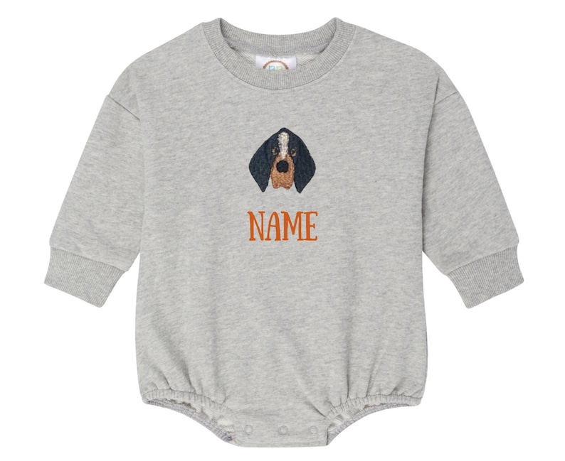 May include: A pale grey baby romper with long sleeves and a round neckline. The romper has an embroidered dog face with black, brown, and white details. The word "NAME" is embroidered in orange below the dog's face.