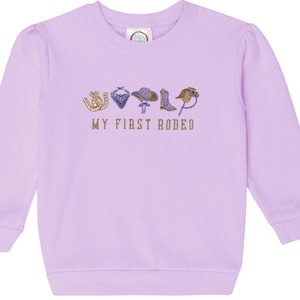Może przedstawiać: Jasnofioletowa bluza z długimi rękawami i złotym napisem "MY FIRST RODEO". Haft przedstawia podkowę, chustę, kapelusz, but i lasso. Bluza ma okrągły dekolt i bufiaste rękawy.