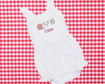 May include: White baby romper with a football theme. The romper features a cartoon dog, a football helmet, and a football. The name "Clara" is embroidered in a cursive font. The romper has ruffled edges and a bubble silhouette.