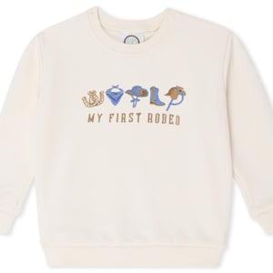 Może przedstawiać: Kremowa bluza z haftowanym napisem "MY FIRST RODEO" i grafiką w stylu rodeo. Projekt zawiera podkowę, bandanę, kapelusz kowbojski, but i lasso. Top z długim rękawem jest odpowiedni dla dzieci.