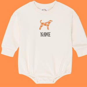 May include: A cream-coloured baby bodysuit with long sleeves. It features an orange and white plaid dog graphic and the word "NAME" in black. The bodysuit has snap closures at the bottom and a round neckline.