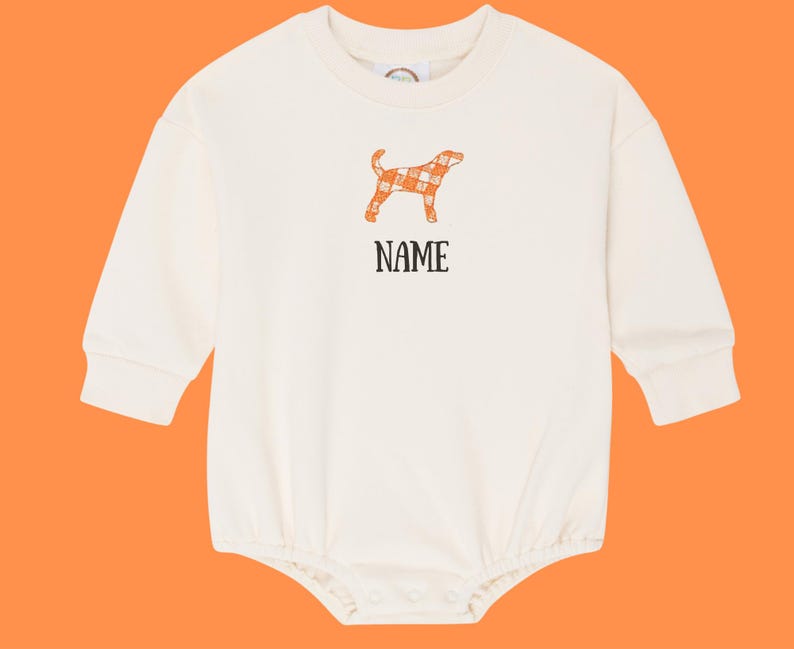 May include: A cream-coloured baby bodysuit with long sleeves. It features an orange and white plaid dog graphic and the word "NAME" in black. The bodysuit has snap closures at the bottom and a round neckline.