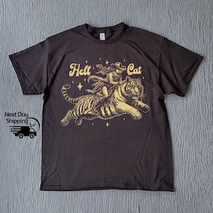 Könnte beinhalten: Schwarzes T-Shirt mit einem goldenen Aufdruck einer Frau, die auf einem Tiger reitet. Der Aufdruck enthält den Text "Hell Cat" und Sterne.