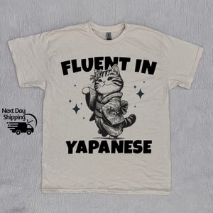 Pode incluir: Uma camiseta bege com um gráfico preto de um gato vestindo um quimono e o texto "Fluent in Japanese".