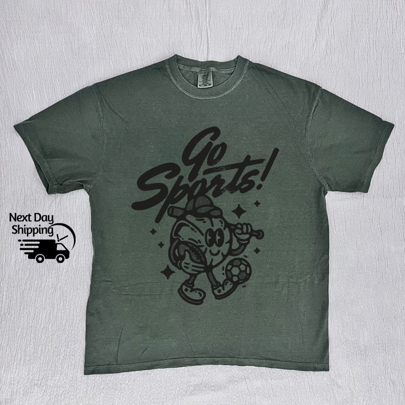 May include: A dark green t-shirt with a black graphic of a cartoon character playing sports. The character is holding a baseball bat and a soccer ball. The text "Go Sports!" is written in a stylized font above the character.