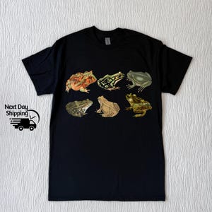 Puede incluir: Camiseta negra con un estampado gr&aacute;fico de seis ranas diferentes en varios colores y poses.