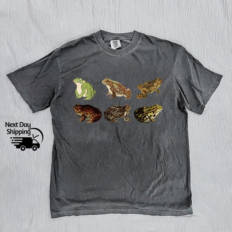 May include: A dark gray t-shirt with a graphic print of six frogs in different poses. The frogs are green, brown, and black.