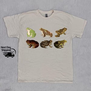 May include: A light gray t-shirt with a graphic print of six different frogs in various poses. The frogs are in shades of green, brown, and black.