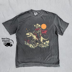 May include: Dark grey t-shirt with a skeleton cowboy riding a dinosaur under a full moon. The cowboy wears a hat and red scarf. The graphic is set against a starry sky and cloud background.