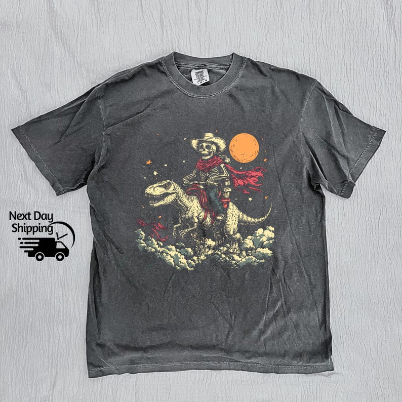 May include: Dark grey t-shirt with a skeleton cowboy riding a dinosaur under a full moon. The cowboy wears a hat and red scarf. The graphic is set against a starry sky and cloud background.