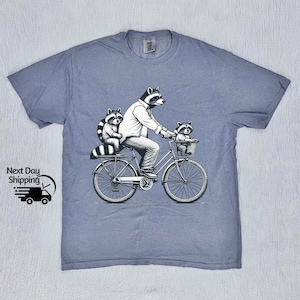 May include: A light blue t-shirt with a graphic of three raccoons riding a bicycle. The raccoon in the front is wearing a shirt and riding the bike. The two raccoons in the back are sitting in a basket.