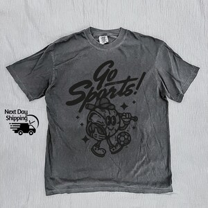 May include: A dark gray t-shirt with a black graphic of a cartoon sports mascot holding a baseball bat and a soccer ball. The text "Go Sports!" is above the mascot.