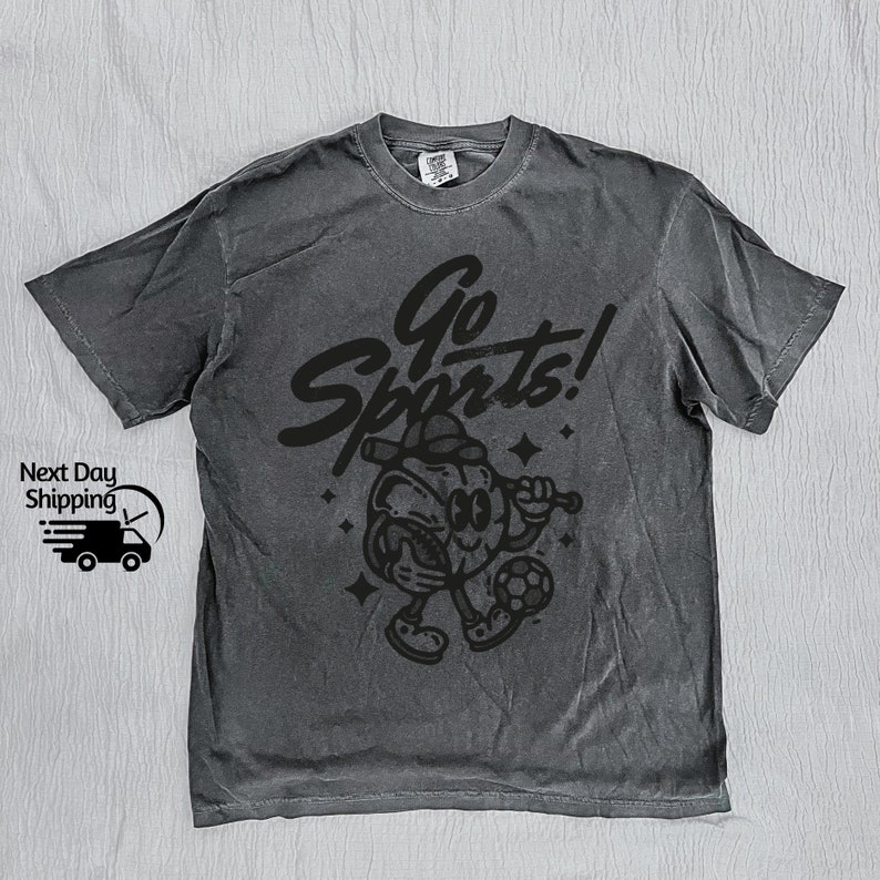 May include: A dark gray t-shirt with a black graphic of a cartoon sports mascot holding a baseball bat and a soccer ball. The text "Go Sports!" is above the mascot.