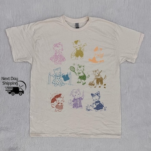 K&ouml;nnte beinhalten: Ein cremefarbenes T-Shirt mit einem Aufdruck von verschiedenen Katzen in verschiedenen Posen und Farben. Die Katzen tragen Kleidung und halten Gegenst&auml;nde wie einen Tennisschl&auml;ger, eine Gie&szlig;kanne und einen Eimer.