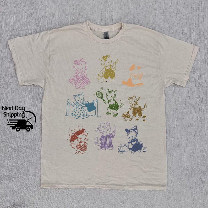 K&ouml;nnte beinhalten: Ein cremefarbenes T-Shirt mit einem Aufdruck von verschiedenen Katzen in verschiedenen Posen und Farben. Die Katzen tragen Kleidung und halten Gegenst&auml;nde wie einen Tennisschl&auml;ger, eine Gie&szlig;kanne und einen Eimer.