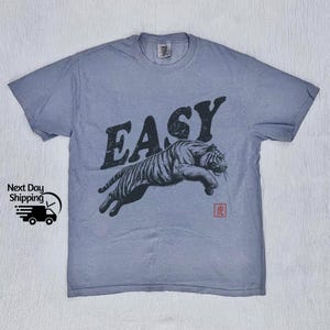 K&ouml;nnte beinhalten: Ein hellblaues T-Shirt mit einem springenden schwarzen Tiger und dem Wort "EASY" in gro&szlig;en schwarzen Buchstaben. Das Wort "虎" ist in Rot unter dem Tiger gedruckt.