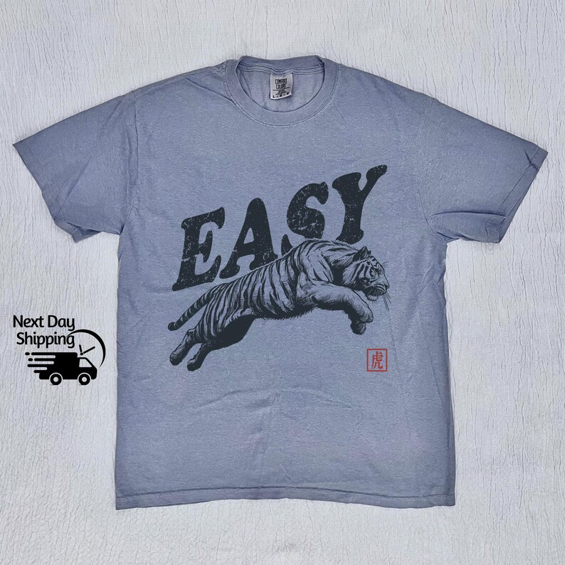 K&ouml;nnte beinhalten: Ein hellblaues T-Shirt mit einem springenden schwarzen Tiger und dem Wort "EASY" in gro&szlig;en schwarzen Buchstaben. Das Wort "虎" ist in Rot unter dem Tiger gedruckt.