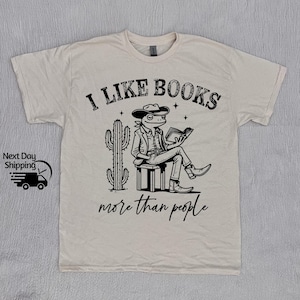 May include: A beige t-shirt with a black and white graphic of a frog wearing a cowboy hat and reading a book. The text "I like books more than people" is printed on the shirt.