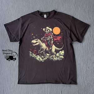 May include: Dark grey t-shirt with a graphic of a skeleton cowboy riding a dinosaur under a full moon. The cowboy wears a hat and red scarf. The graphic is in beige, red, and blue.